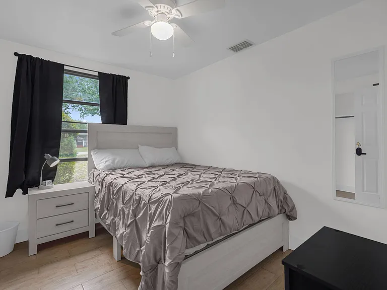 Furnished bedroom with a comfortable bed, nightstand, ceiling fan, and a large window with blackout curtains.