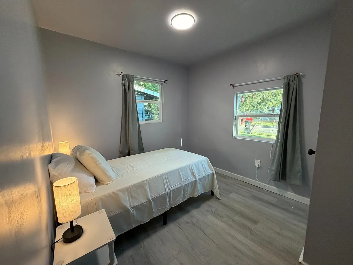 Photo 1 of 7 - Huntington Dr - Arlington, TX Private bedroom with a furnished bed, side table, and two windows with curtains for natural light and airflow. - Huntington Dr