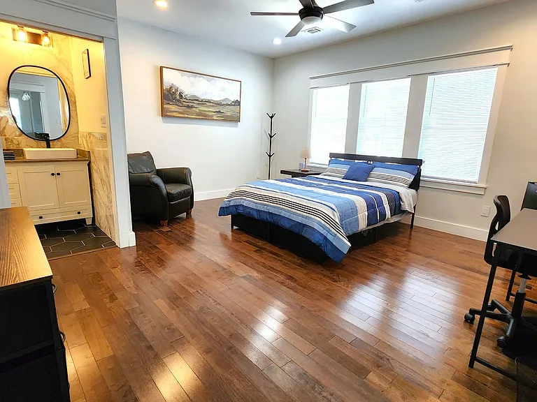 Spacious furnished bedroom with hardwood floors, large windows, ceiling fan, dedicated desk, and a private vanity area.