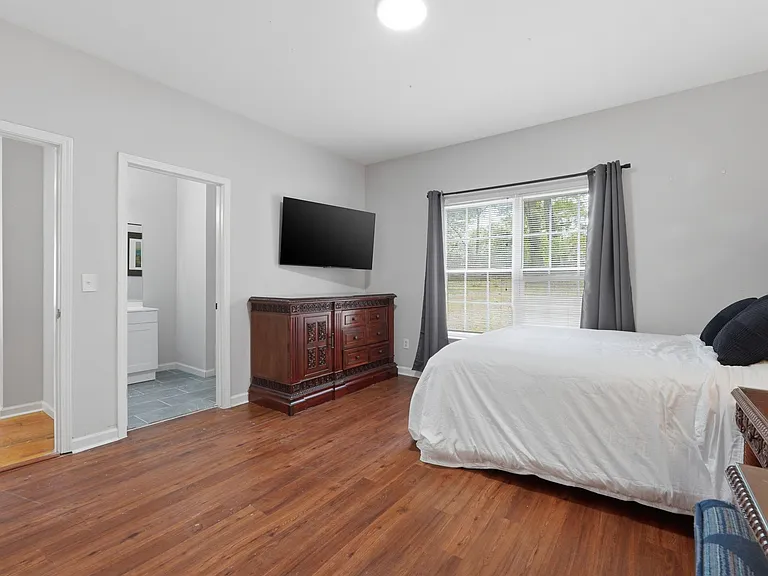 Spacious bedroom with hardwood floors, large window with drapes, dresser storage, and a wall-mounted TV near an en-suite bath
