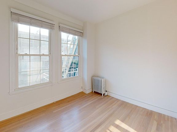 Bright, empty room with hardwood floors, featuring two large windows on the left. A small radiator sits beneath one window.