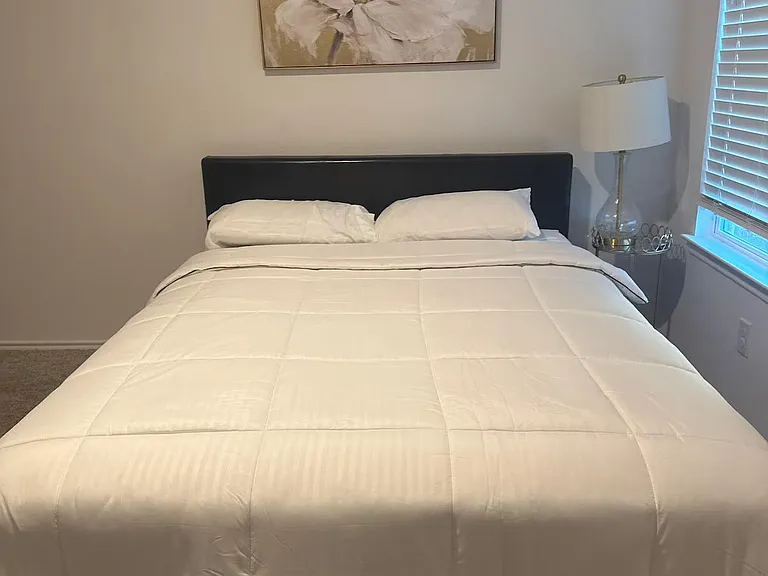 Furnished bedroom with a comfortable bed, bedside table and lamp, and a window with blinds for light and privacy.