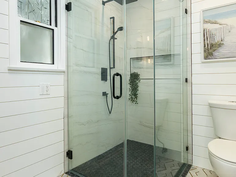 Bright, beautiful bathroom with a spacious glass-enclosed walk-in shower, rainfall and handheld fixtures, a double vanity, designer lighting, shiplap walls, and elegant marble-patterned tile.