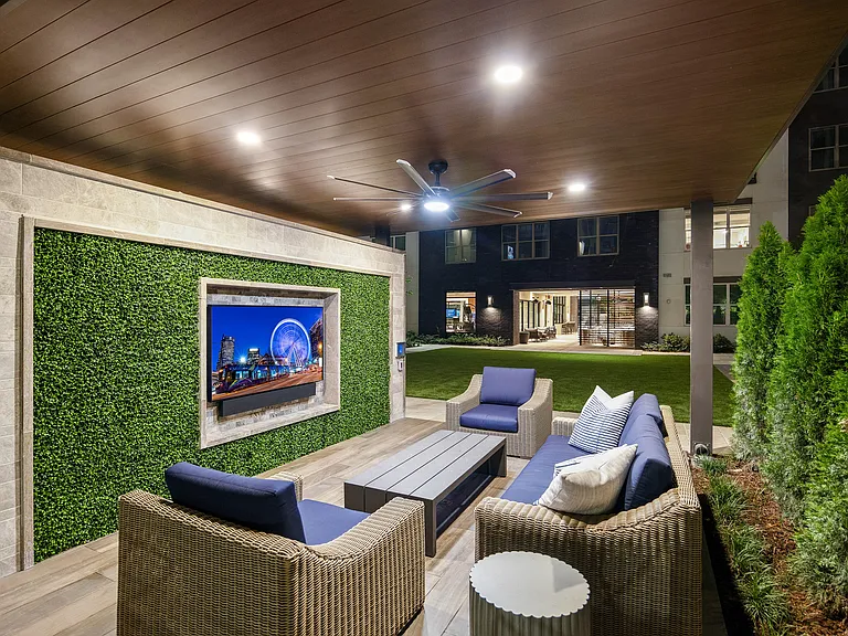 Elegant outdoor lounge area with stylish wicker furniture, modern ceiling fan, and vibrant greenery wall featuring a mounted television. Perfect space for entertaining and relaxation under the night sky.