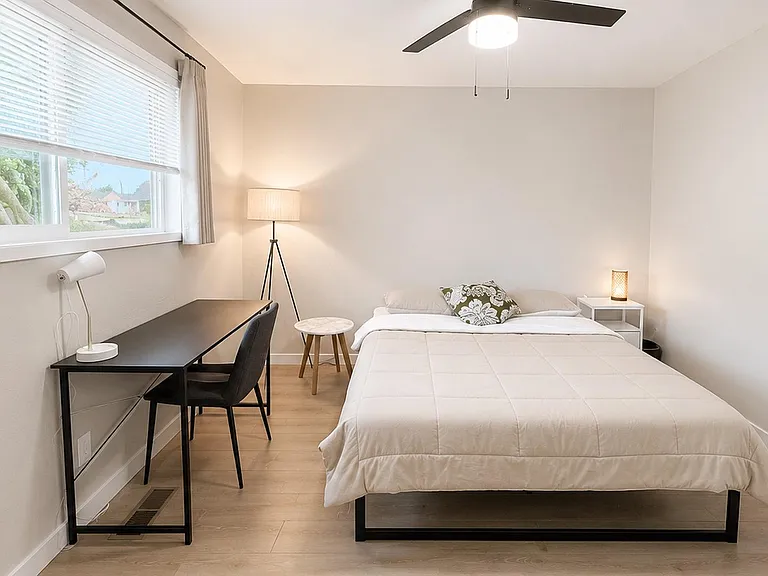 Furnished bedroom with a full bed, dedicated desk workspace, ceiling fan, and a large window with blinds and curtains.