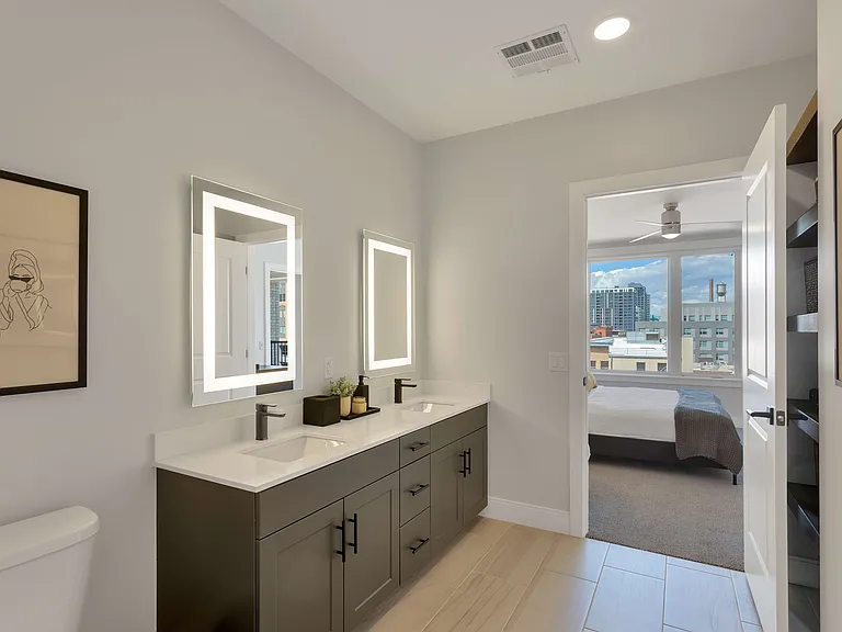 Modern bathroom with dual sinks and illuminated mirrors, flowing into a bright bedroom with large windows offering city views.
