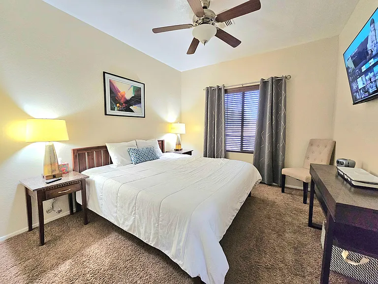Furnished bedroom with queen bed, nightstands, ceiling fan, and a window with blinds and curtains for privacy.
