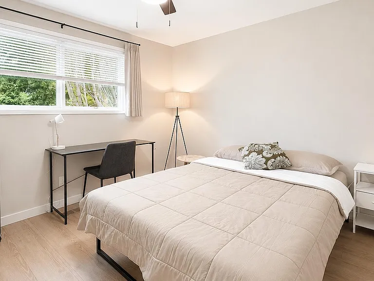 Private bedroom with a full bed, bedside table, and dedicated desk workspace, plus a large window with curtains.