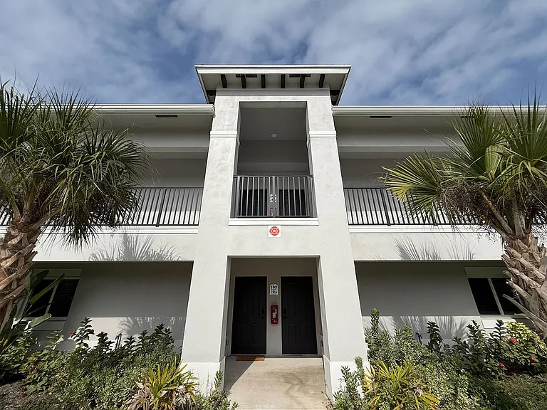 Modern apartment exterior with lush landscaping and palm trees, featuring a clean white facade and elegant balcony.