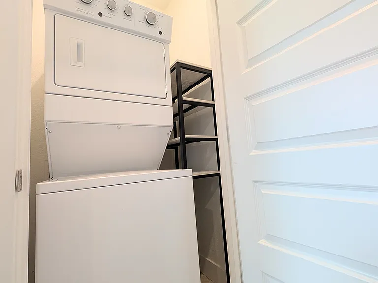 Convenient in-unit laundry area equipped with a stacked washer and dryer, plus a tall multi-tier storage rack to keep your laundry supplies organized and easily accessible.