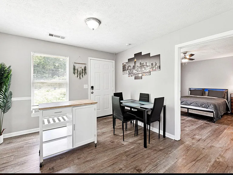 Dining area with a 4-seat table, nearby serving cart, and a large window with blinds; open to adjacent bedroom.