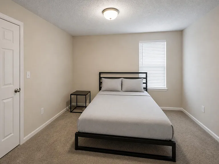 Furnished bedroom with a full bed, nightstand, carpeted flooring, and a window with blinds for privacy and daylight.