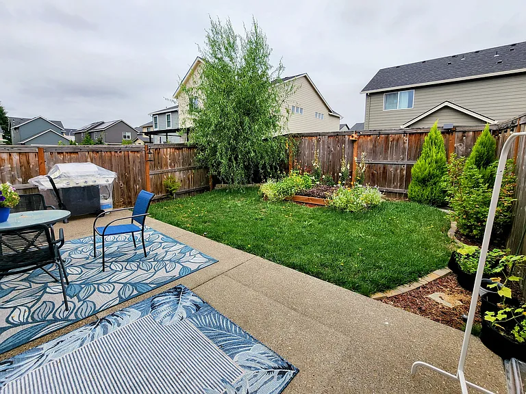 Fully fenced backyard with patio seating area, lush lawn, fully grown trees, and established garden beds