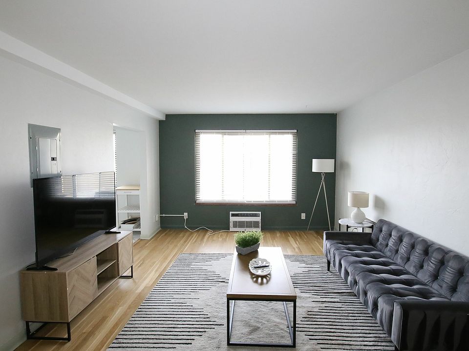 Modern living room with sleek black sofa, large window with blinds and natural light, stylish wooden TV stand, and minimalist decor.
