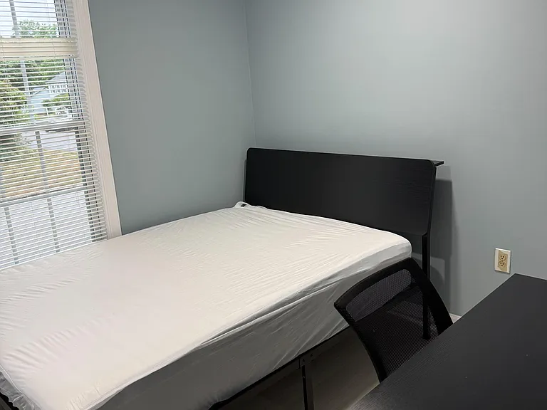 Private bedroom with a full bed, desk workspace, and a large window with blinds for natural light and privacy.