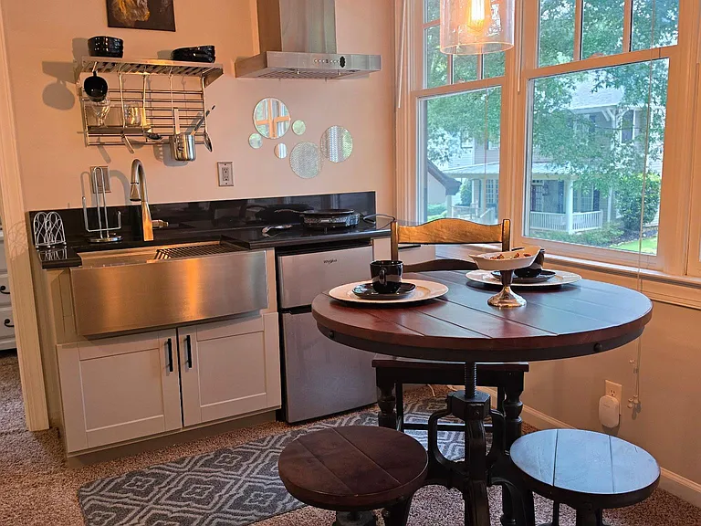 Compact eat-in kitchen with stainless appliances, vent hood, open wall storage, and a round dining table by large windows.