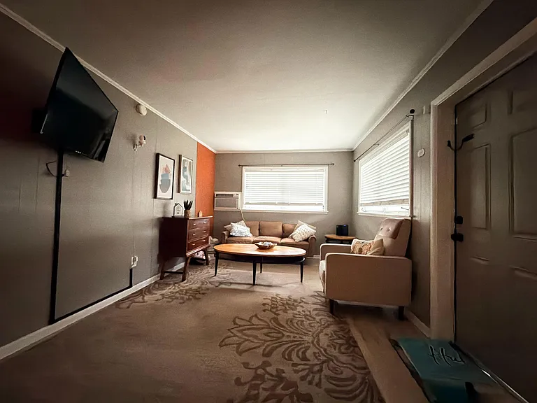 Shared living room with seating area, coffee table, wall-mounted TV, and dual windows with blinds for natural light.