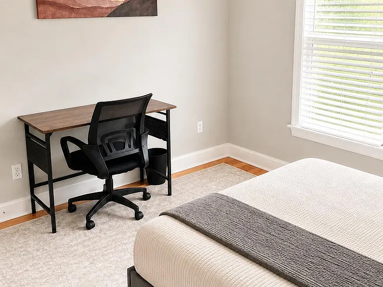 Well-appointed bedroom with a comfortable queen bed, dedicated desk, and a large window with blinds for natural light.
