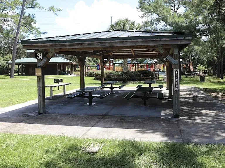 Covered outdoor picnic pavilion with multiple tables, nearby green space, and easy access to walking paths and amenities.