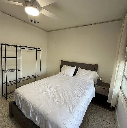 Furnished bedroom with a full bed, nightstand, and open shelving, plus a window with curtains for natural light.