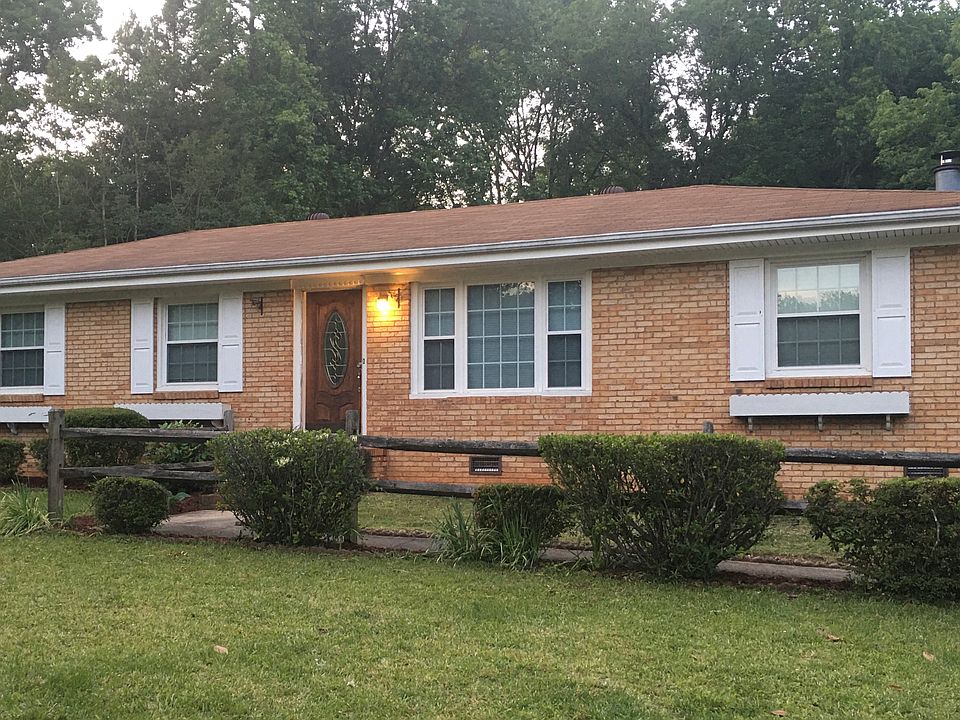 Front of the house featuring a split rail fence lining the walkway, flower beds with bushes, three Pomas bushes, and two additional trees (one small and one large).