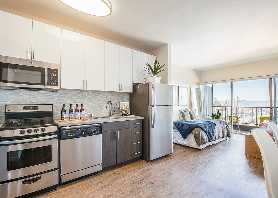 Staged studio with hardwood style flooring, sliding balcony doors, and kitchen with stainless steel appliances.