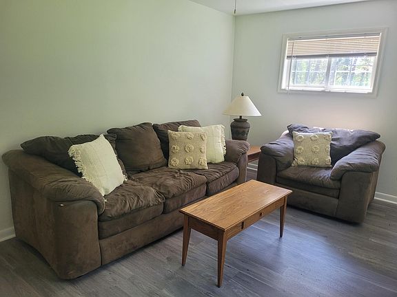 Living room with comfortable couch, overstuffed chair tables, lamp and more. View out window is wooded.