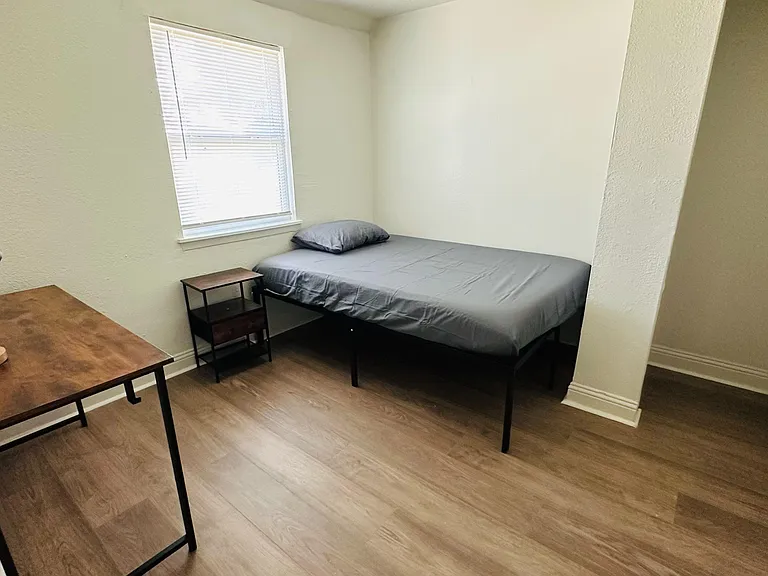 Furnished bedroom with a comfortable bed, side table, and dedicated desk workspace, plus a window with blinds.