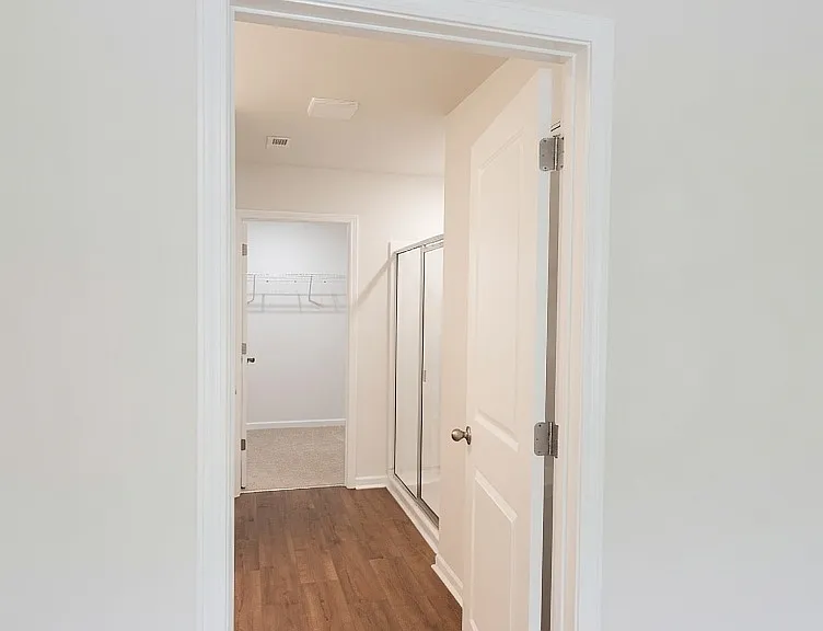View into the primary bathroom corridor showing the glass shower enclosure, walk-in closet beyond, and clean wood-look flooring. Soft lighting and white finishes create a spa-like feel.