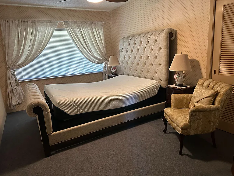Spacious furnished bedroom featuring a tufted bed, side tables with lamps, and a large window with drapery for natural light.