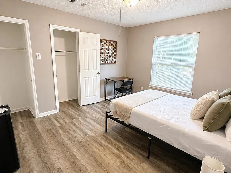Furnished bedroom with wood-look flooring, large window, open closet storage, and a compact desk area for work or study.