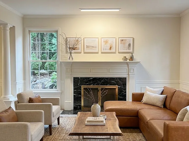 Bright formal living area boasting expansive windows, wainscoting, and a centerpiece fireplace with marble surround and gold accents. (computer staged)