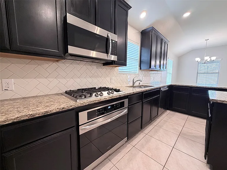 Modern kitchen featuring dark cabinetry, stainless steel appliances, speckled stone countertops, and a herringbone tile
 backsplash