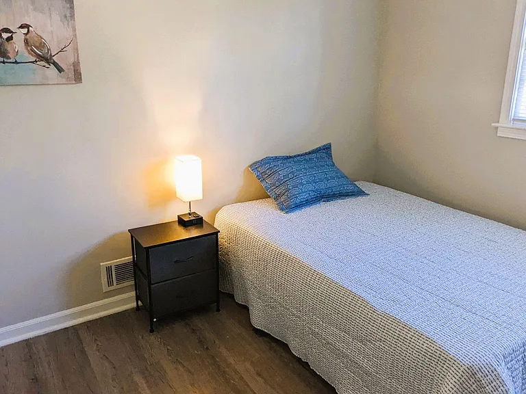 Furnished bedroom with wood-look flooring, a twin bed, nightstand with lamp, and a window with blinds for natural light.