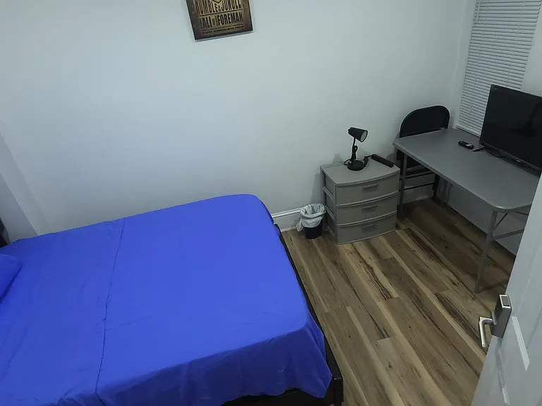 Private bedroom features a full bed, desk setup with TV, and wood-style flooring with closet storage nearby.