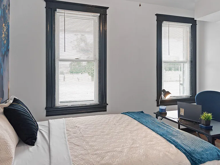 Bedroom with a full bed, two large windows with blinds, and a compact desk setup for work or study.