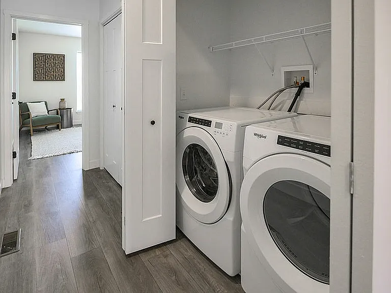 Modern laundry area with sleek washer and dryer units, complemented by elegant wood flooring and a glimpse into a cozy, stylish room.