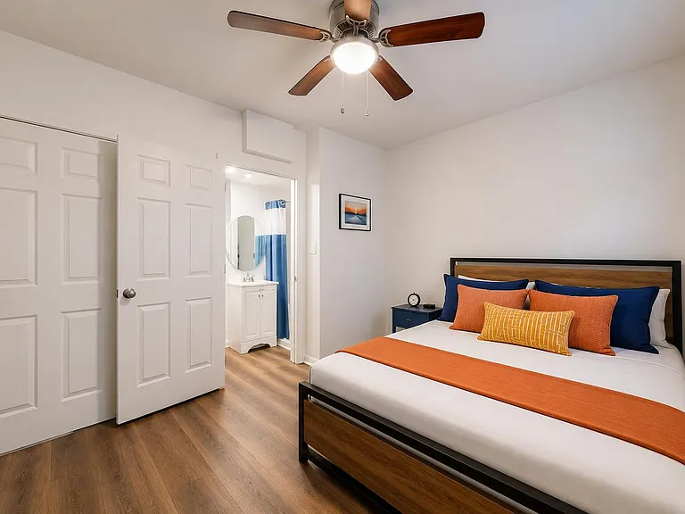 Furnished bedroom with a ceiling fan, large window with blinds, and easy access to an adjacent bathroom vanity area.
