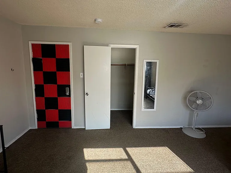 Carpeted bedroom with a reach-in closet and space for a bed and desk, plus overhead lighting and a standing fan.