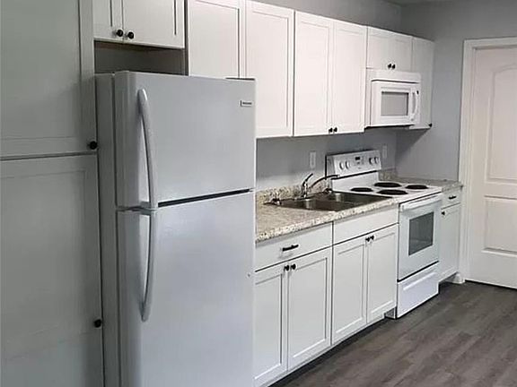 Kitchen with white appliances, sink, white cabinets, light stone countertops, and dark wood-type flooring