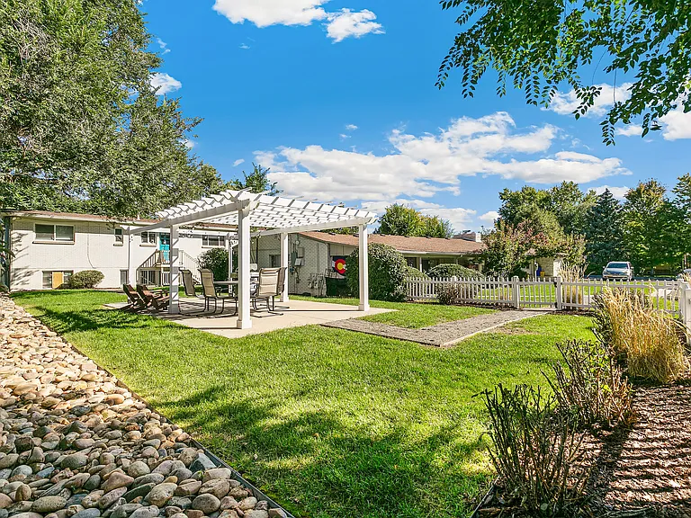Charming backyard with a pergola and seating area, surrounded by lush greenery and a white picket fence under a bright blue sky.