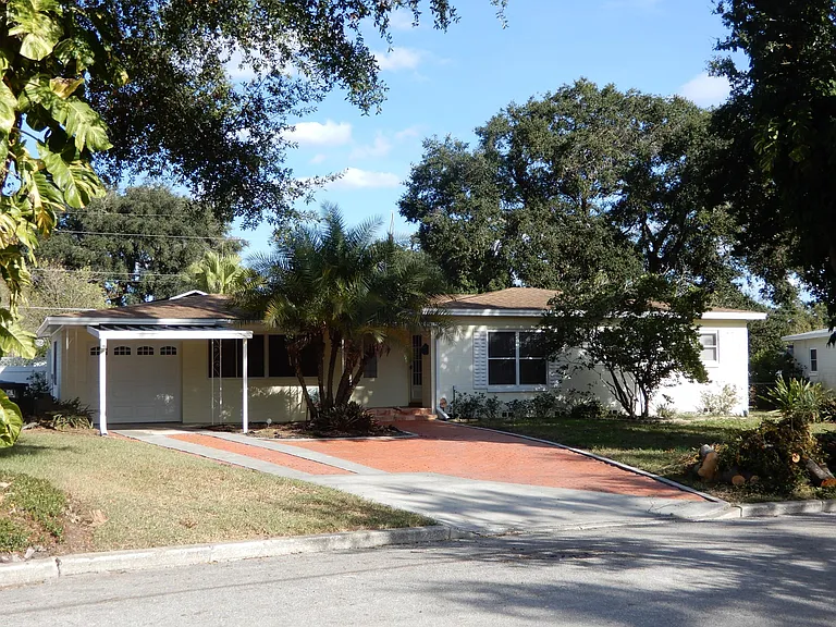 Frontal view of house facing east shows red clay brick parking area with the ability to park 3 vehicles across when necessary. The single car garage has a metal roof extension to keep your vehicles out of the sun and rain.