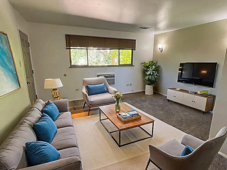 Inviting living room with a modern gray sofa, blue accent pillows, and cozy armchairs. Large window offers natural light over a stylish rug and elegant coffee table.