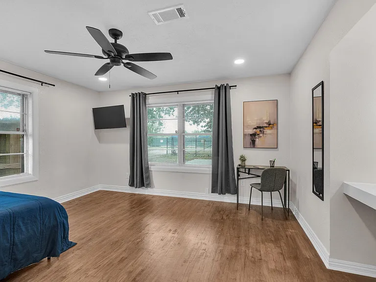 Spacious bedroom with wood-look floors, ceiling fan, and two windows with blinds and curtains plus a work desk area.