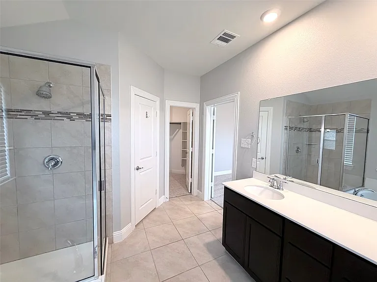 Spa-inspired primary bathroom featuring a spacious walk-in shower with frameless glass enclosure and tile surround, dual-sink vanity with dark cabinetry and white quartz countertop, large framed mirror with integrated lighting, and neutral tile flooring t