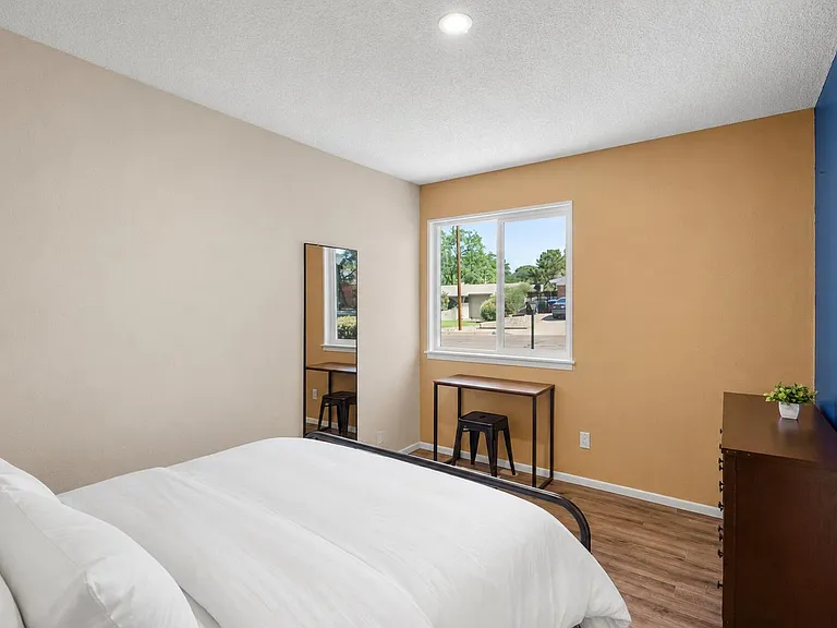 Furnished bedroom with a large window, dedicated desk/vanity area, full-length mirror, and dresser for extra storage.