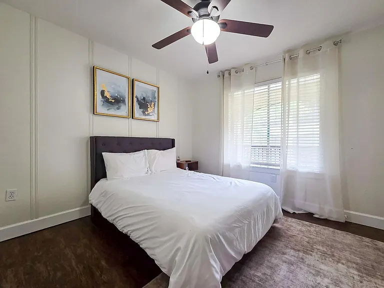 Bedroom with a comfortable bed, ceiling fan light, and large window with blinds and curtains for natural light and privacy.
