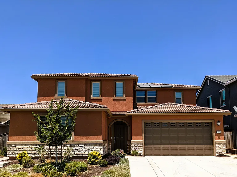 -Modern two-story home with a stucco exterior and tiled roof. Features a three-bay garage, arched entryway, and multiple windows.
-Landscaped front yard with various shrubs and plants, set in a quiet residential neighborhood.