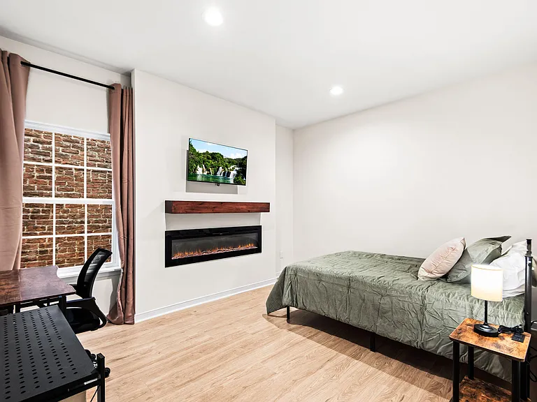 Private furnished bedroom with desk workspace, wall-mounted TV, and an electric fireplace; includes window and nightstand.