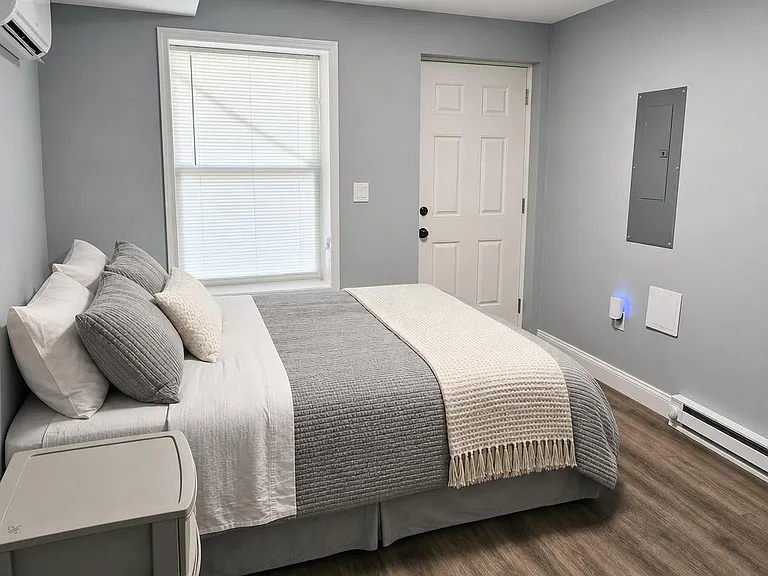 Furnished bedroom with a comfortable bed, window with blinds, and in-room A/C for added comfort.